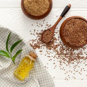 Flaxseed is scattered on a table. There are bowls and a spoon full of flaxseed. There's also a small bottle of linseed oil next to some green leaves. 