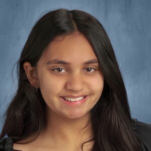 A young woman with long dark hair and brown eyes smiles softly at the camera. She is wearing a black top and small hoop earrings, with a blue textured background behind her.