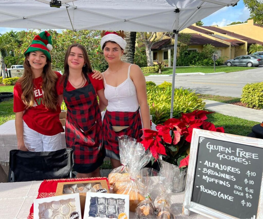 Emi, Lulu and Raffa in red Christmas-themed clothes stand outside behind a table. Various baked goods are visible, as well as a menu showing the gluten-free offerings. 