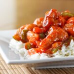 A close-up of sweet and sour chicken with bell peppers and sauce served over white rice on a white plate, placed on a bamboo mat.
