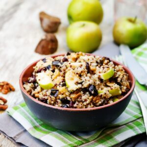 A bowl of quinoa salad with apple slices, dried cranberries, and walnuts sits on a striped cloth, with apples, walnuts, and cutlery visible in the background on a wooden table.