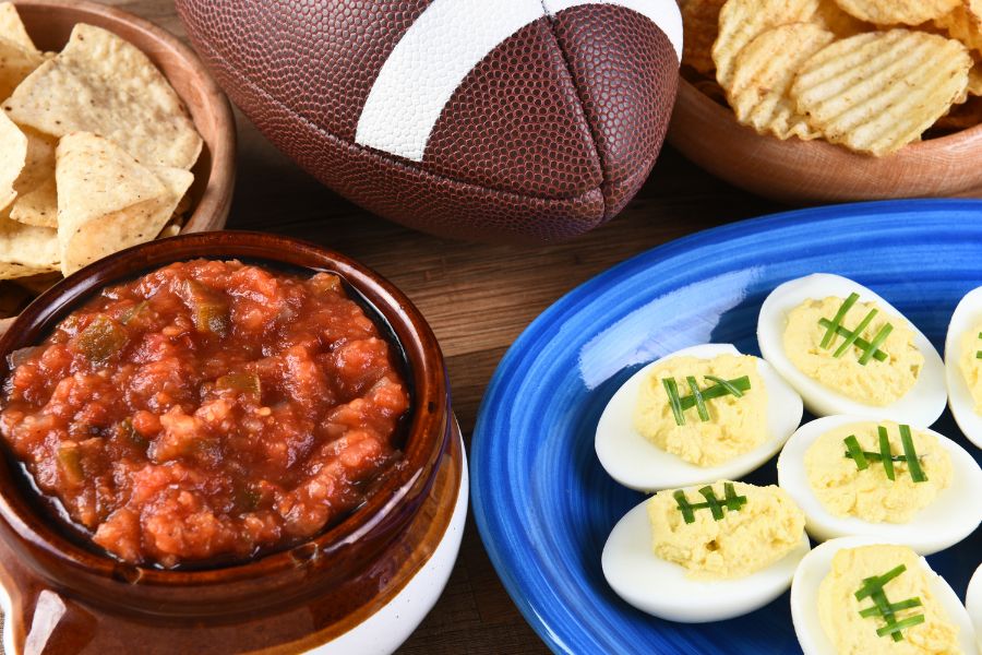 Bowls of tortilla chips, potato chips, salsa, and deviled eggs on a table next to a football.