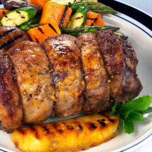A plate with slices of roasted meat, grilled vegetables including zucchini, asparagus, and sweet potato, a piece of grilled pineapple, and a sprig of mint as garnish.