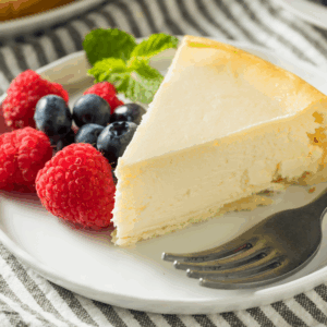 A slice of plain cheesecake on a white plate with fresh raspberries, blueberries, and a sprig of mint, next to a fork on a striped cloth.