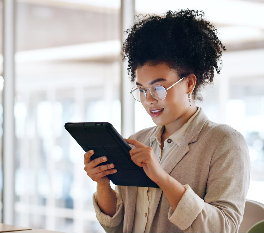 A woman wearing glasses and a light-colored blazer is sitting indoors, looking at and using a tablet device. Bright windows and office furniture are visible in the background.