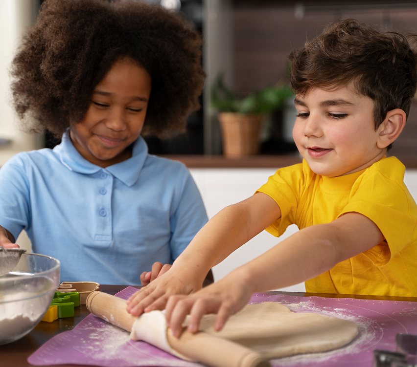 Two children are baking together. One child in a blue shirt smiles while the other, wearing a yellow shirt, uses a rolling pin to flatten dough on a purple mat. Baking tools and flour are visible on the table.