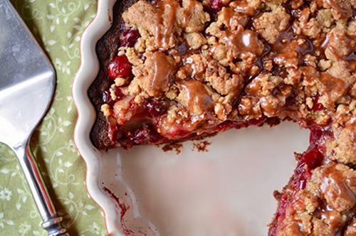 A cherry crumble pie with a slice missing, showing a crumbly topping and juicy filling in a round ceramic pie dish, next to a metal pie server on a green patterned cloth.
