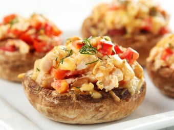 Four stuffed mushrooms are arranged on a white plate. The mushrooms are filled with a mixture containing chopped vegetables and herbs, and topped with melted cheese. The filling appears colorful and slightly browned.