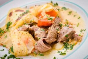 A plate of sliced cooked meat with thin potato slices, carrots, and a light sauce, garnished with chopped parsley.