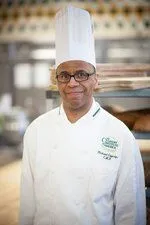 A chef wearing a tall white hat and a white chef coat stands in a kitchen with metal racks and baked goods in the background.
