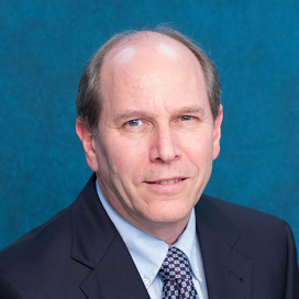 A middle-aged man with short, thinning hair is wearing a dark suit, light blue shirt, and patterned tie, and is posing in front of a plain blue background.