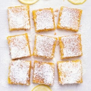 Nine square lemon bars topped with powdered sugar are arranged in a grid on a white surface, with a few lemon slices partially visible around the edges.