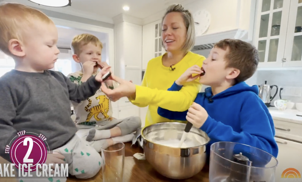 A woman and three young boys sit around a kitchen counter. The woman holds a cookie while one boy licks it, another boy eats a cookie, and a third boy watches. There is a mixing bowl and measuring cups on the counter.