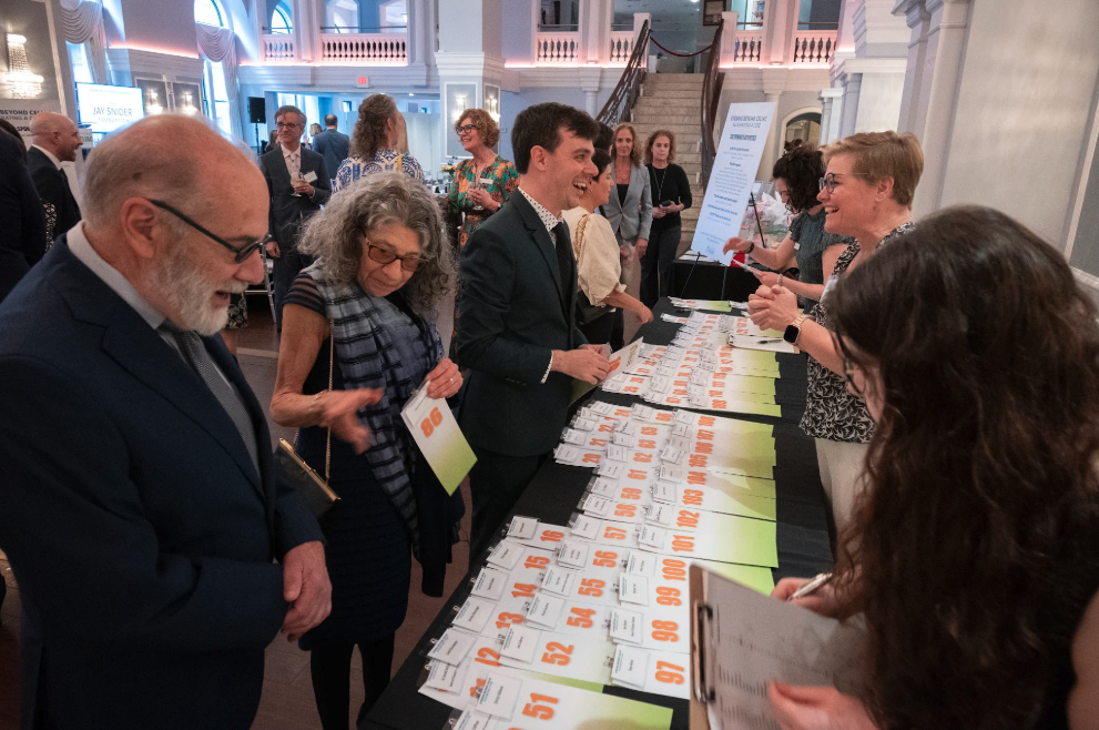 People check in at a registration table covered with numbered badges at a formal event. Attendees are dressed in business or semi-formal attire inside a spacious, well-lit venue.