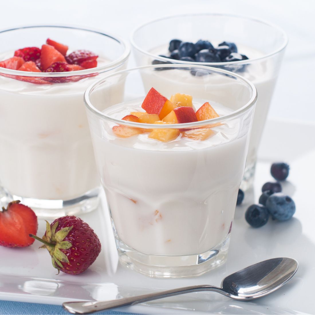 Three glasses of yogurt topped with chopped fresh fruit, including strawberries, blueberries, and peach pieces, are arranged on a white tray next to a spoon and some loose berries.