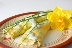 Two rolled crepes with a creamy herb sauce on top, garnished with fresh chives, are served on a plate next to a yellow daffodil flower.