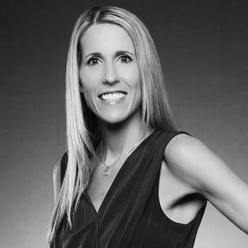 A woman with long straight hair, wearing a sleeveless top and a necklace, stands with one hand on her hip, smiling at the camera. The image is black and white with a plain background.