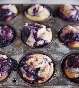 A close-up of a metal muffin tin holding nine baked blueberry muffins. The muffins have a marbled appearance with purple blueberry swirls on top and some batter is slightly overflowing in a few cups.