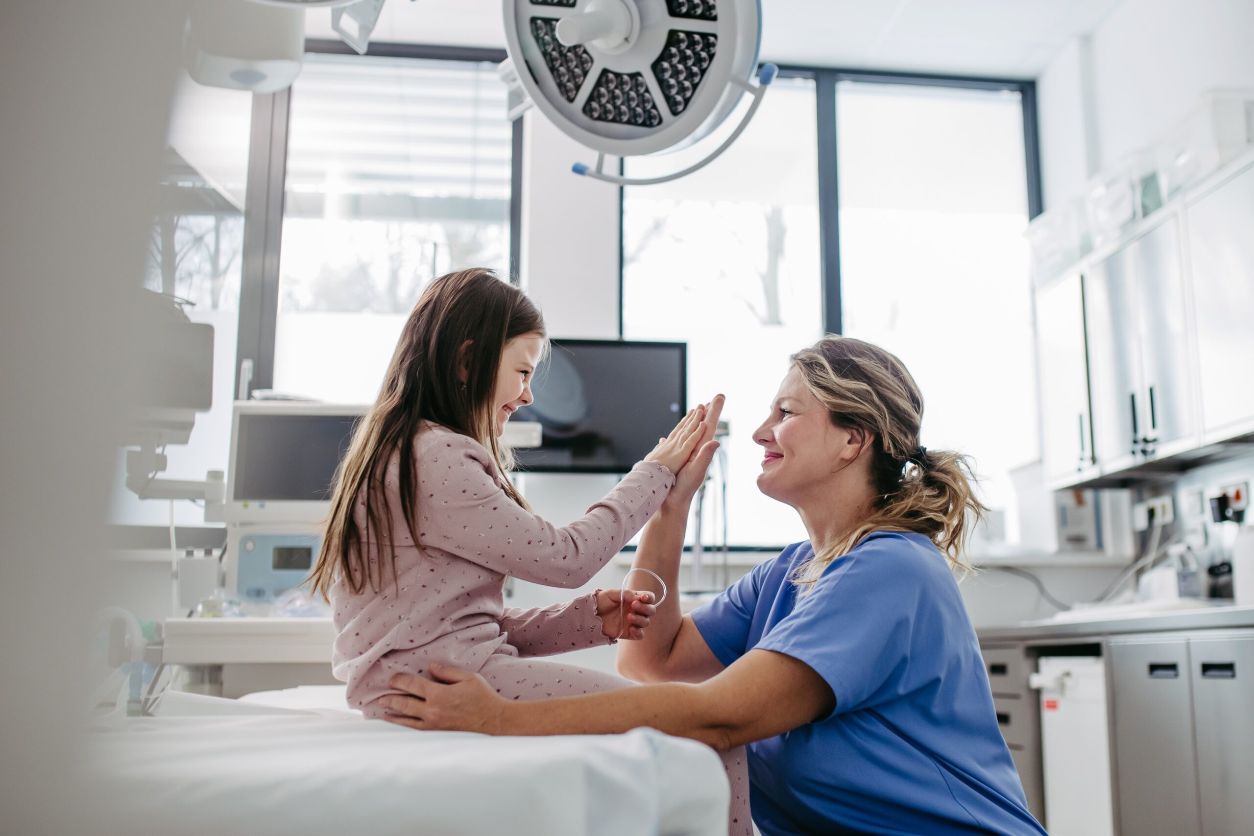 A young girl in pajamas sits on an exam bed giving a high-five to a smiling healthcare worker in blue scrubs in a bright, modern medical room.