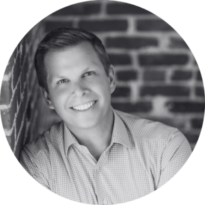 A man with short hair, wearing a checked shirt, leans against a brick wall and smiles at the camera. The image is in black and white and cropped in a circular frame.