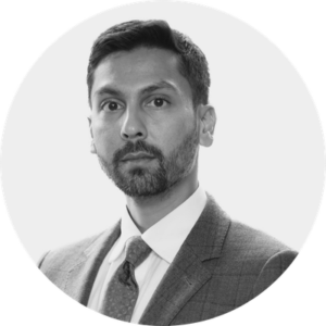 A man with short dark hair and a beard, wearing a checked suit jacket, white shirt, and tie, looks directly at the camera against a plain light background. The black and white image reflects professionalism—ideal for beyond celiac investments branding.