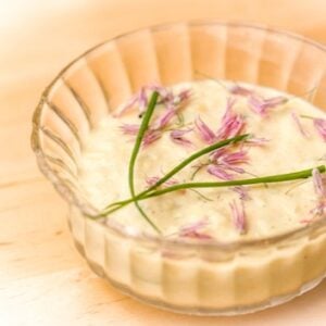 A small glass bowl filled with creamy dip, garnished with chive stems and purple flower petals, sits on a light wooden surface.