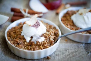 Two white ramekins filled with apple crumble topped with a scoop of vanilla ice cream and a thin apple slice sit on burlap fabric. A spoon rests in one ramekin. Cinnamon sticks and apple slices are in the background.