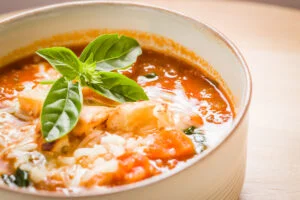 A bowl of tomato soup topped with shredded cheese, toasted bread pieces, and a fresh basil sprig, served in a light-colored bowl on a wooden surface.