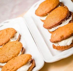Two white trays hold several sandwich cookies with golden-brown cakes and a thick layer of white cream filling. The trays are placed on a light-colored surface, and the cookies are arranged neatly in rows.