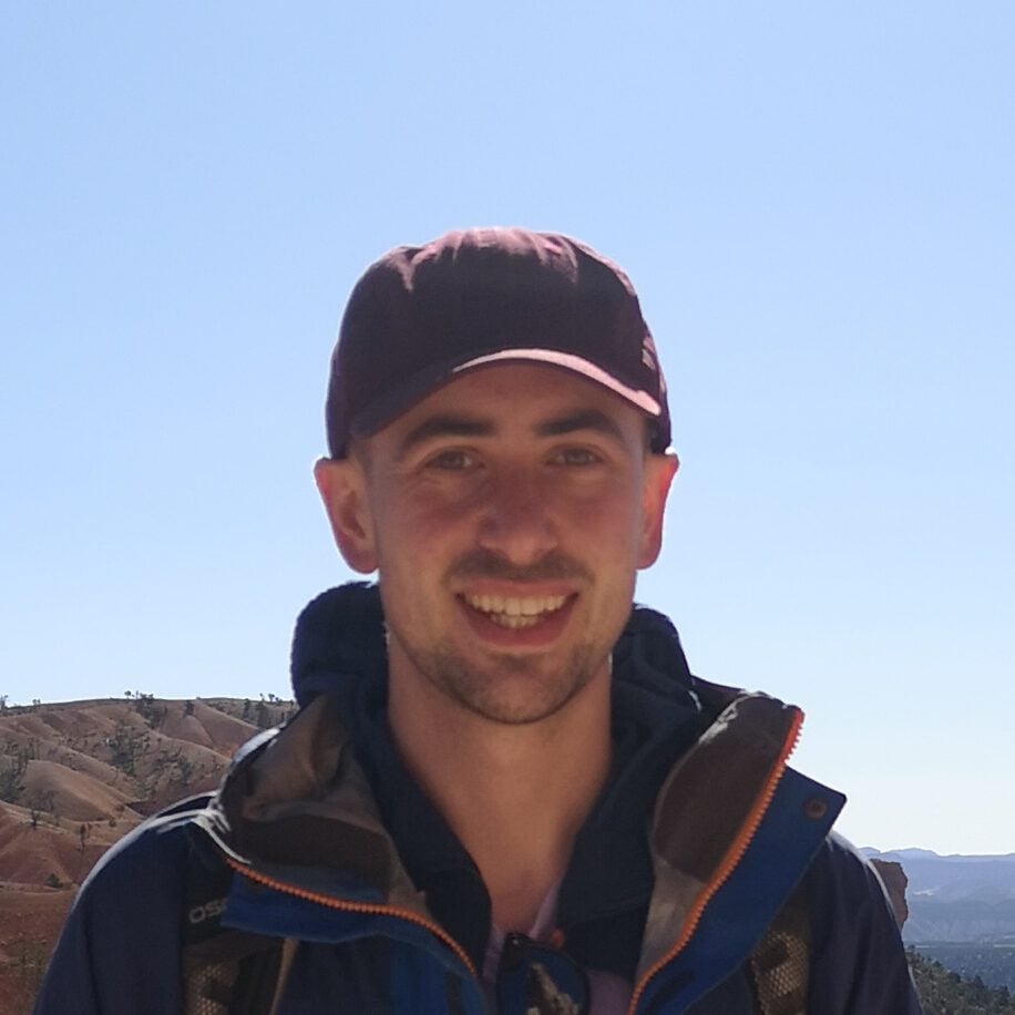 A smiling man wearing a baseball cap and a blue hiking jacket stands on a desert trail in Bryce Canyon.